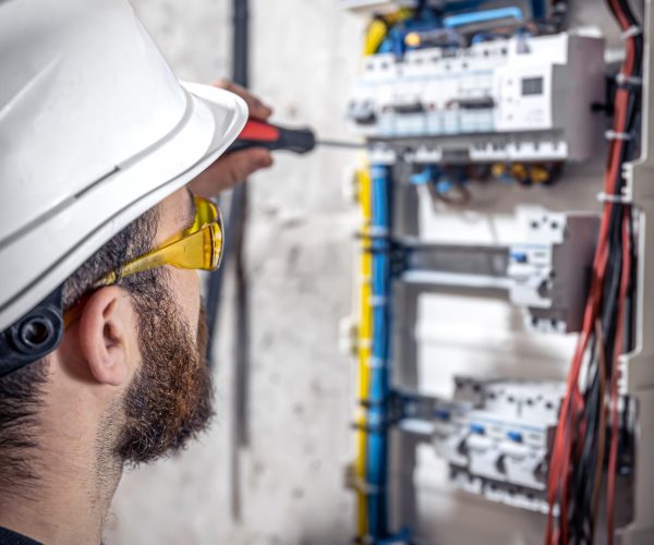 A male electrician works in a switchboard with an electrical connecting cable, connects the equipment with tools.