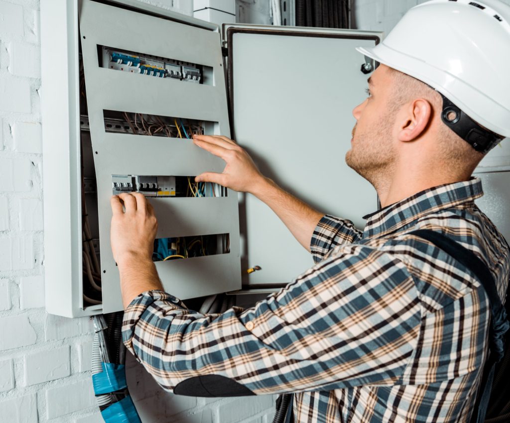 electrician in safety helmet looking at switchboard near brick wall